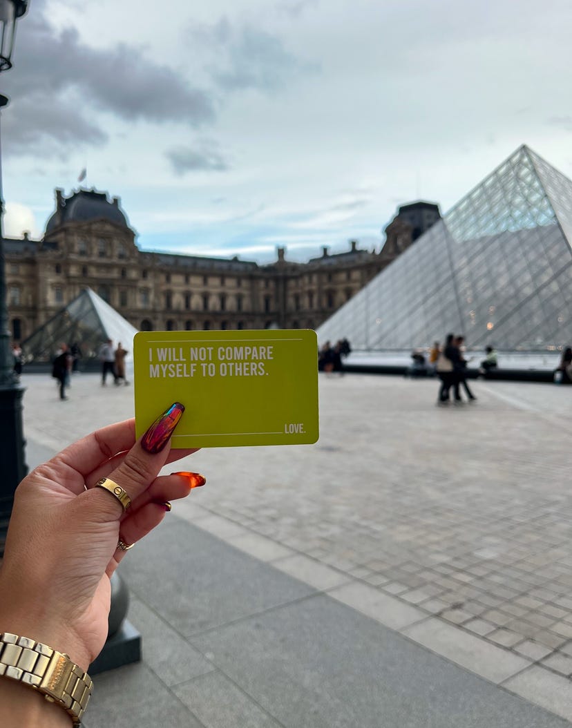 An affirmation card saying I will not compare myself to others being held up between two glass pyramids at the Louvre.