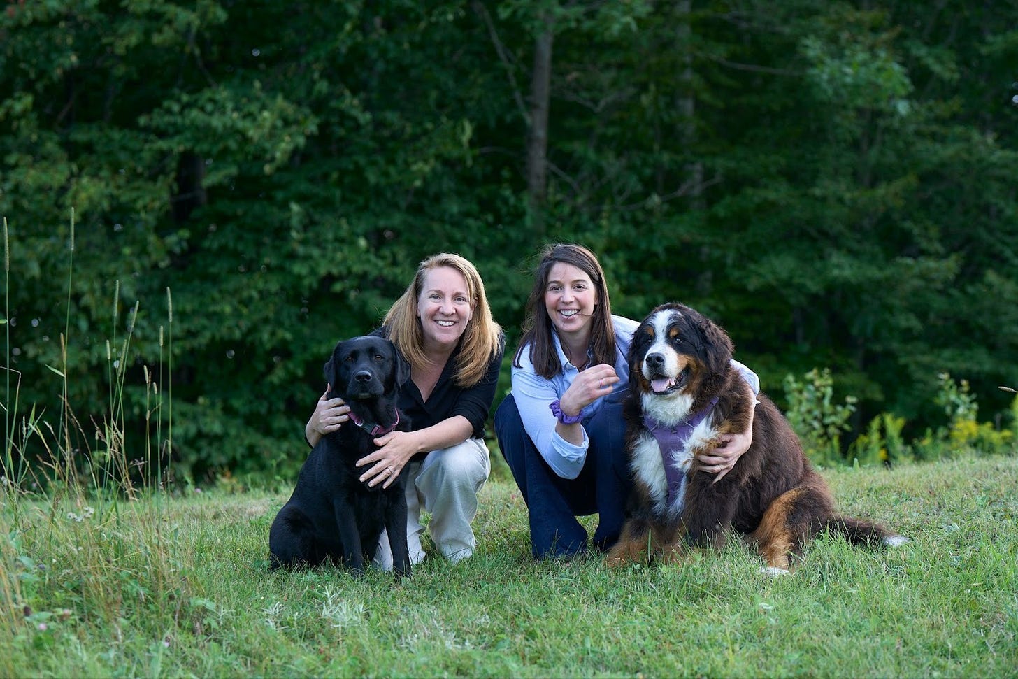 Amy and Kate crouch in a wooded field in Vermont, each holding one of Kate's two service dogs and smiling. Amy and Kate crouch in a wooded field in Vermont, each holding one of Kate's two service dogs and smiling.