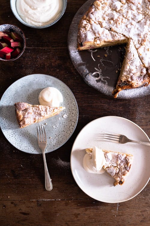 Rhabarber Kuchen mit Streuseln und Sahne. Zwei Stücke sind aus dem Kuchen heraus geschnitten und liegen jeweils auf ein einem Kuchenteller. Ein Klecks Sahne befindet sich neben jedem Kuchenstück. 