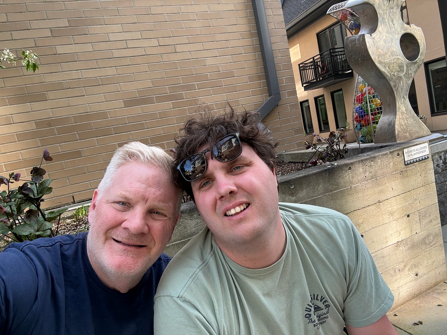 J.B. Handley and his son Jamie smiling cheek to cheek outdoors beside a stone sculpture