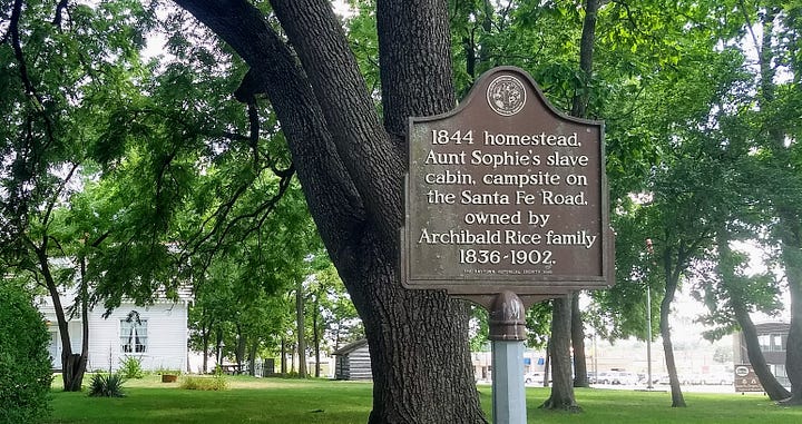 Modest one story house with attic rooms set in large lawn with cars and development glimpsable across the main road plus historic signs