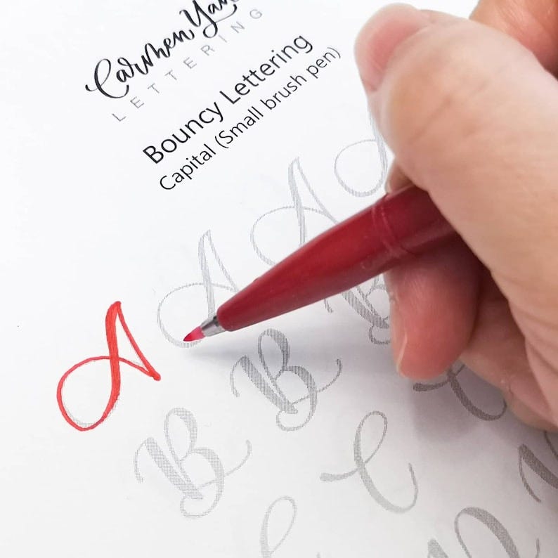 May include: A hand holding a red brush pen, writing the letter "A" in red ink on a white sheet of paper. The paper has the text "Bouncy Lettering Capital (Small brush pen)" printed on it.