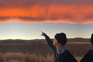 A gallery of photos of Andrea Gibson, an non-binary person, sitting in bed eating from a bowl with her adorable dogs staring at them, walking in a meadow with a dog over their shoulder, staring into a mirror, walking down an empty highway, pointing to a stunning sunrise, and seated at a table. There's also an image they created of them talking to themselves and a photo of their arms crossed with tattoos on their forearms.