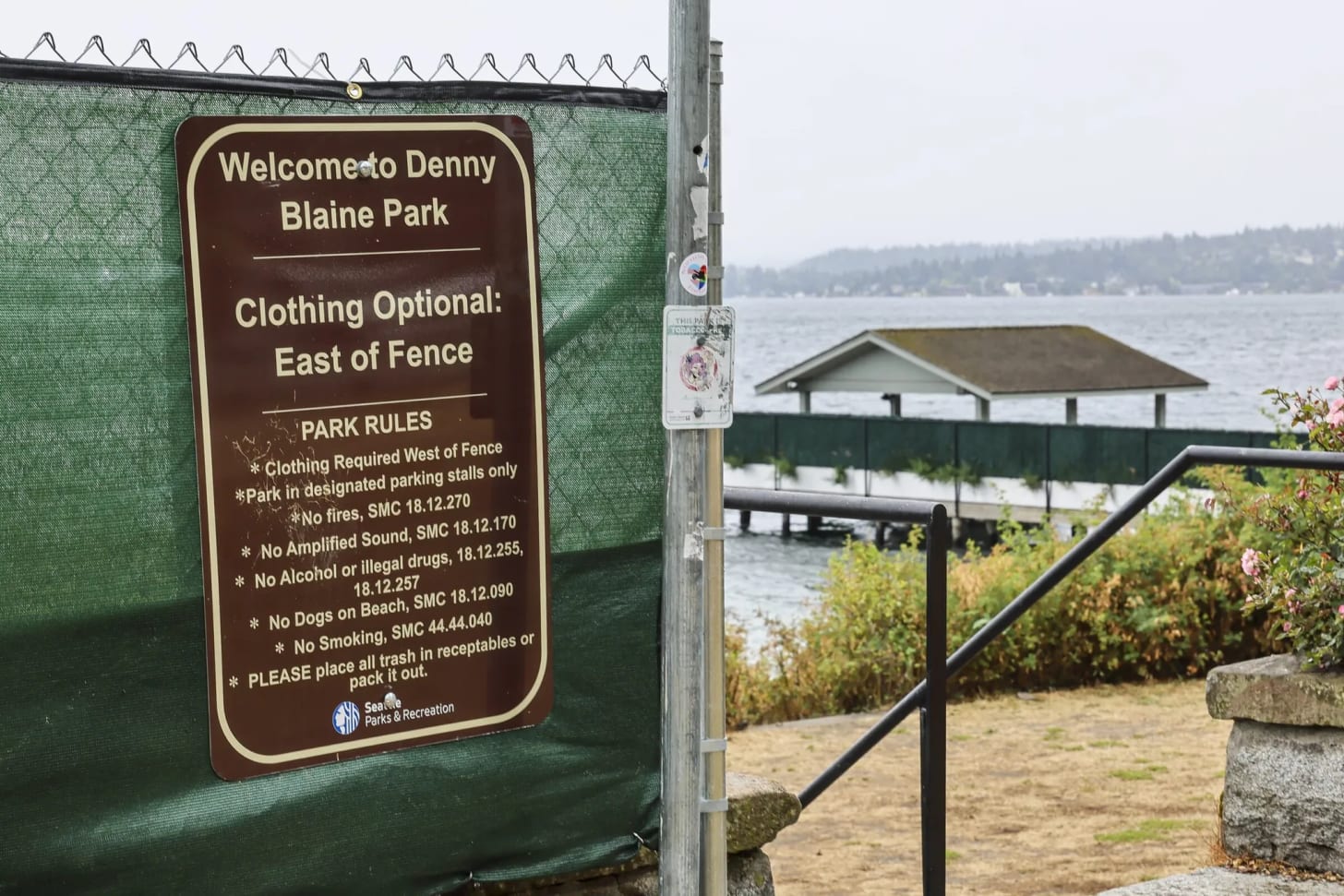 A brown park sign mounted on a chain-link fence wrapped in green privacy mesh reads "Welcome to Denny Blaine Park — Clothing Optional: East of Fence," with park rules listed below including "Clothing Required West of Fence." Lake Washington and a covered dock are visible in the background.