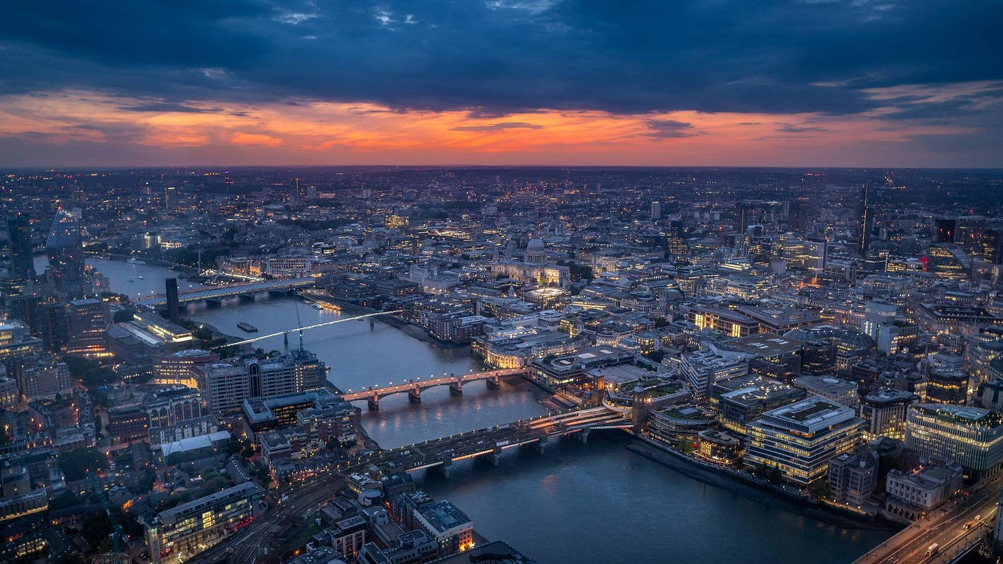 Aerial view of London at dusk with the Thames winding through the illuminated cityscape. St Paul's Cathedral is visible in the centre, with multiple bridges spanning the river. Dark blue skies contrast with a band of orange sunset and the glowing lights of buildings below.