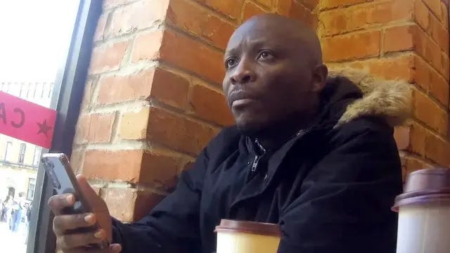 Nana Akwasi Agyemang-Prempeh wearing a navy parka coat with a fur collar, holds his phone in a coffee shop. Nana Akwasi Agyemang-Prempeh wearing a navy parka coat with a fur collar, holds his phone in a coffee shop.