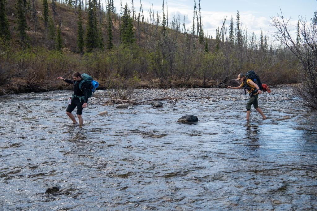 Two hikers with packs cross a wide, braided gravel-bottomed stream from opposite directions, arms out for balance in the moving water, a spruce-covered hillside rising behind.