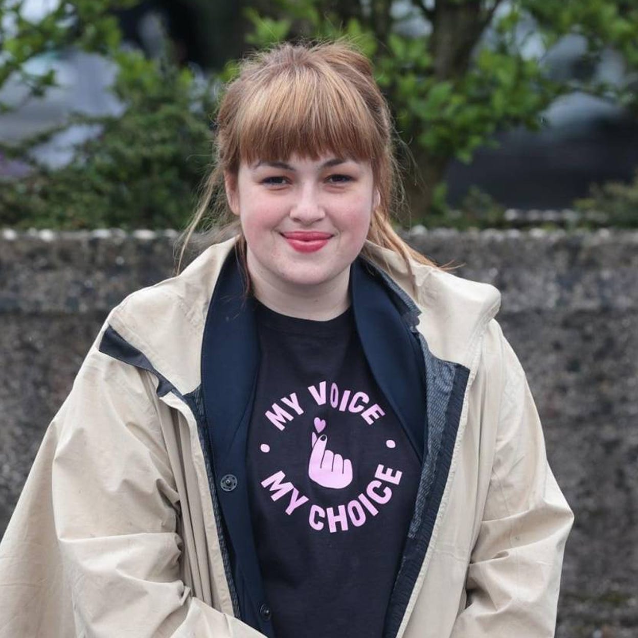Nika Kovač, a white woman with a light skin tone, holds a closed-lip smile. She is wearing a tan jacket and a black shirt that reads “My Voice, My Choice.” She has brown hair and bangs and is wearing pink lipstick. 