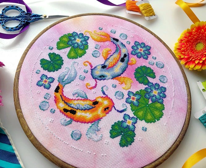 A hooped cross stitch on mottled pink fabric, featuring two koi swirling about each other with lilypads, bubbles, and florals.  The koi are each distinct, one in orange hues with black spots and the other is in bluish white, with orange fins and black spots.