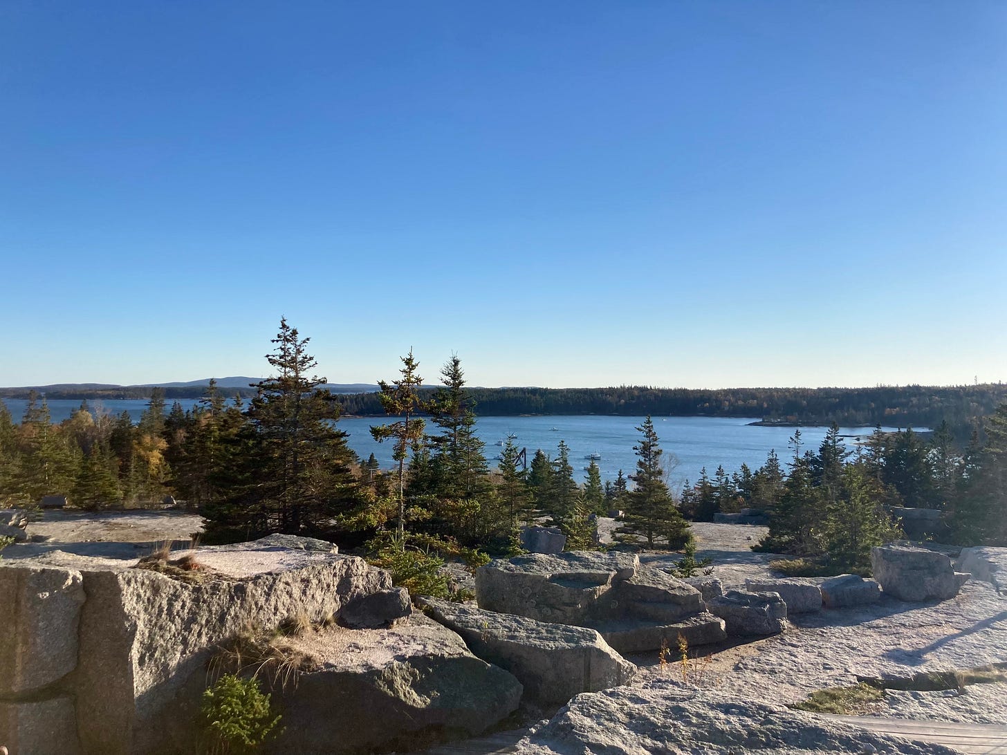 A large excavated area of a granite quarry, with ocean, boats, and cloudless sky in the background. There are evergreens growing throughout the quarry in between the rocks.
