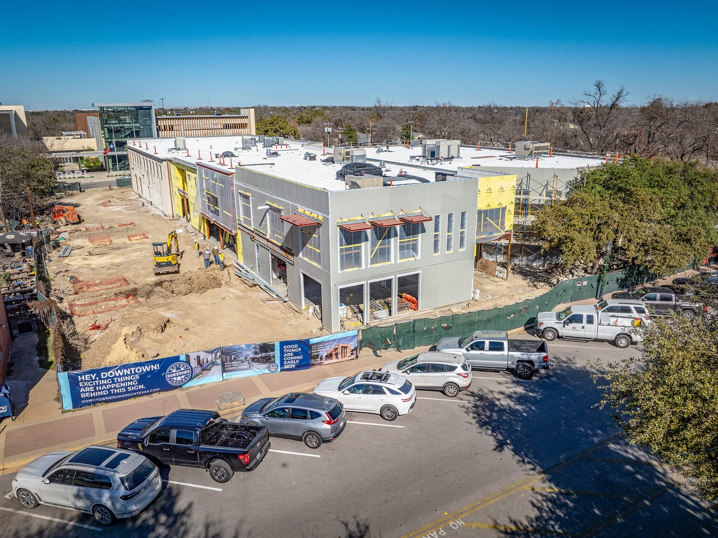 Construction site in downtown Round Rock with a partially completed two-story building. Crews and equipment are visible in the dirt lot, and signage on the fencing reads, “Hey, Downtown! Exciting things are happening behind this sign.” Parked cars line the street in front of the site.