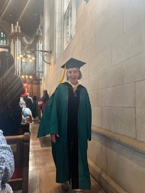 image of older white woman wearing a green graduation cap and gown. She is in a chapel-like building with brick walls and candles hanging from the wall in the background. image of older white woman wearing a green graduation cap and gown. She is in a chapel-like building with brick walls and candles hanging from the wall in the background.