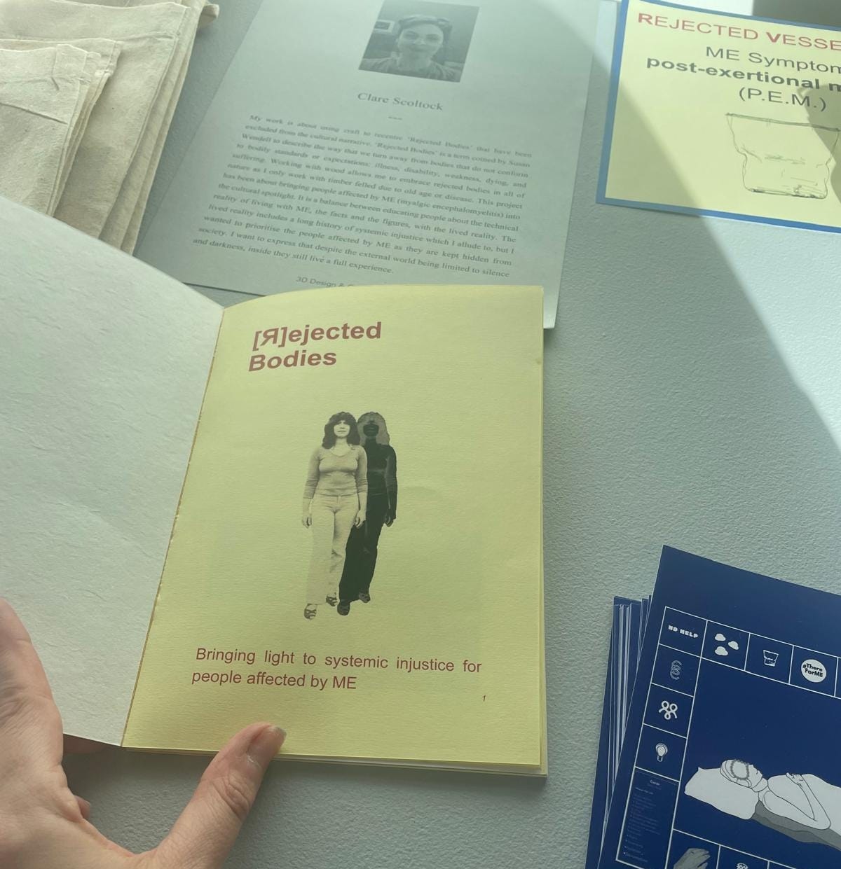 Image is a photo of a booklet at a graduate art fair. The title says "Rejected Bodies" and the subtitle says "Bringing light to systemic injustice for people affected by ME." Next to it are some other less distinct artefacts from this installation.