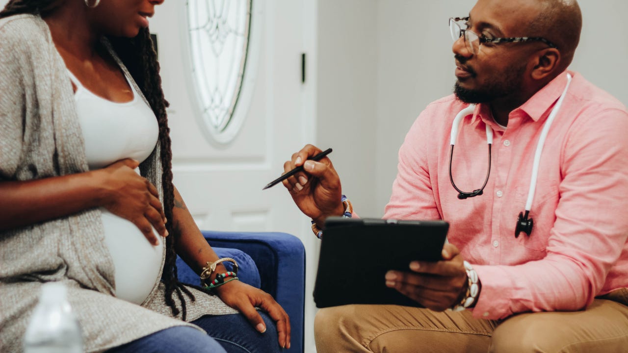 A pregnant Black woman sits in a medical office speaking with a healthcare provider, who holds a tablet and gestures while listening attentively during a prenatal appointment.