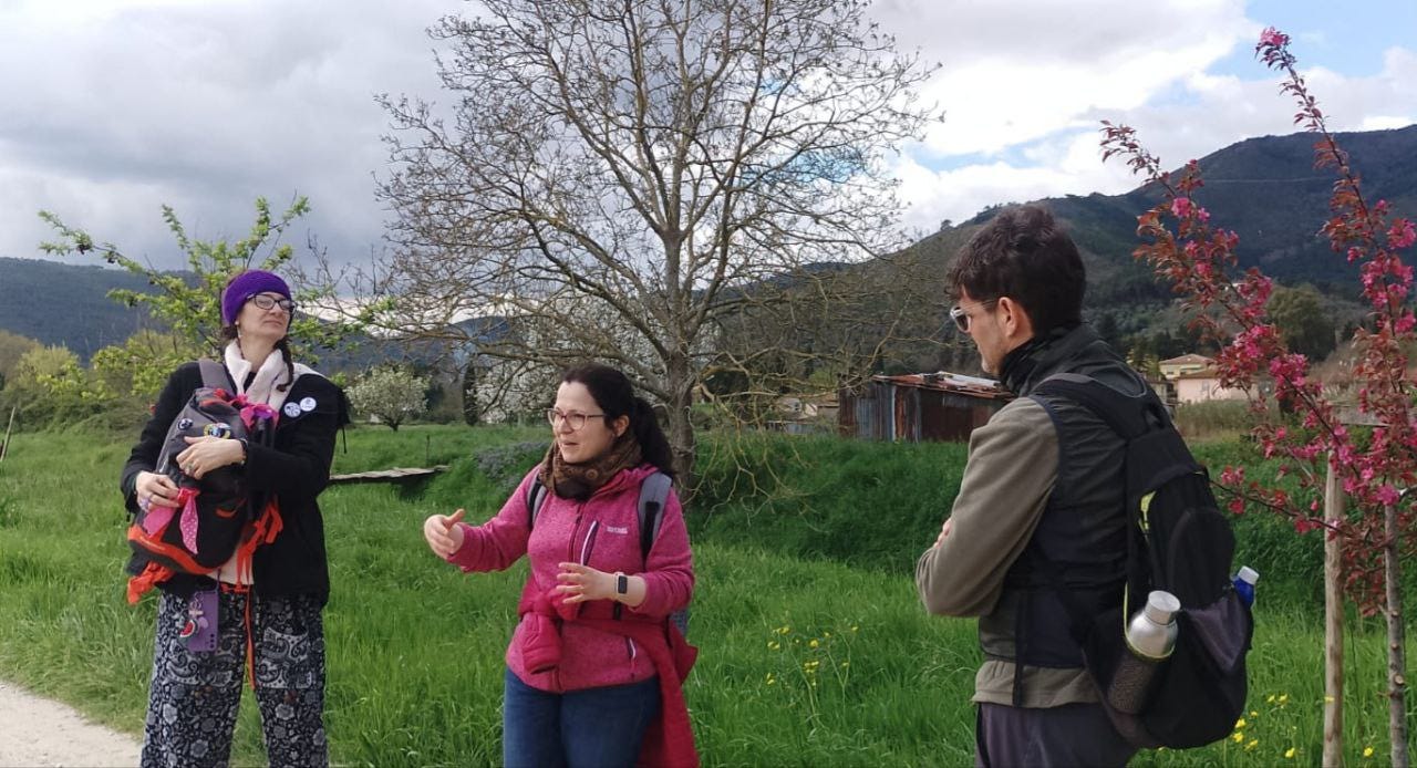 Tre persone sono immerse in un paesaggio naturale con prati verdi, alberi fioriti e colline sullo sfondo. Il cielo è nuvoloso, e l'atmosfera suggerisce un'escursione o un'attività all'aperto.