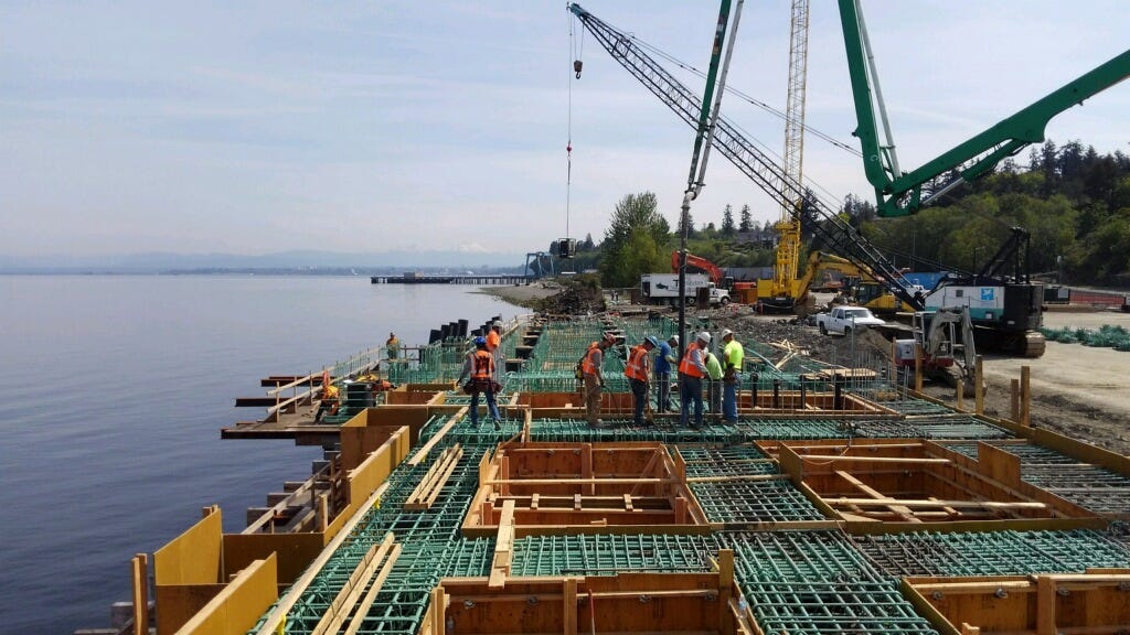 Form works for foundations laid out for a large construction project. A dozen construction workers are walking around, and large cranes are parked nearby.