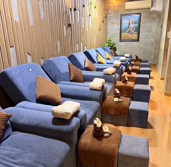 A composite image with a text header that reads, "🧘 Wellness Recommendation: Healing at Comfort Massage." Below the text are two photographs side-by-side. The left photograph shows the exterior of the massage parlor, which has a green facade and a sign that reads "COMFORT M...". It's surrounded by a lush garden with palm trees and shrubs. The right photograph shows the interior foot massage area with a row of blue reclining chairs lined up along a wood-slat wall. Each chair has a footrest with towels, plumeria flowers, and a cluster of stones.