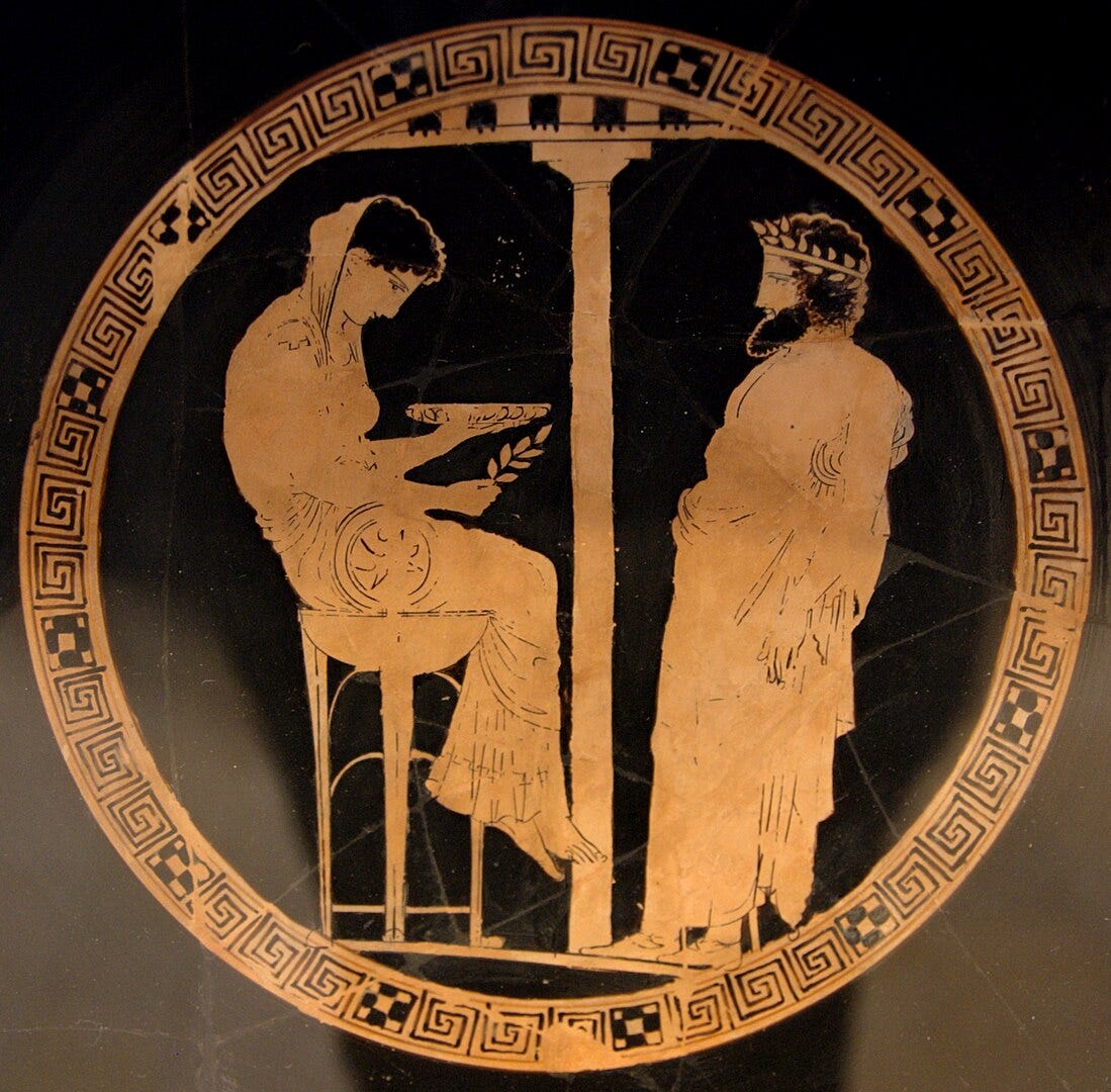 An ancient Greek red-figure kylix interior showing the Pythia, seated on a tripod and holding a laurel branch and bowl, opposite King Aegeus, who wears a crown and long robes. They face each other beneath a central column, encircled by a decorative meander border.