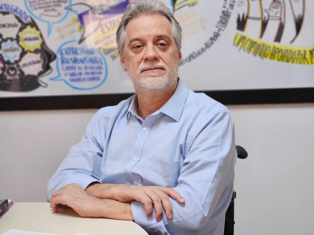 Antonio Claudio é um homem branco, de cabelos grisalhos e barba. Ele usa camisa social azul-clara e está sentado em uma cadeira de rodas, com os braços cruzados sobre a mesa. Ao fundo, parede branca com quadro colorido.