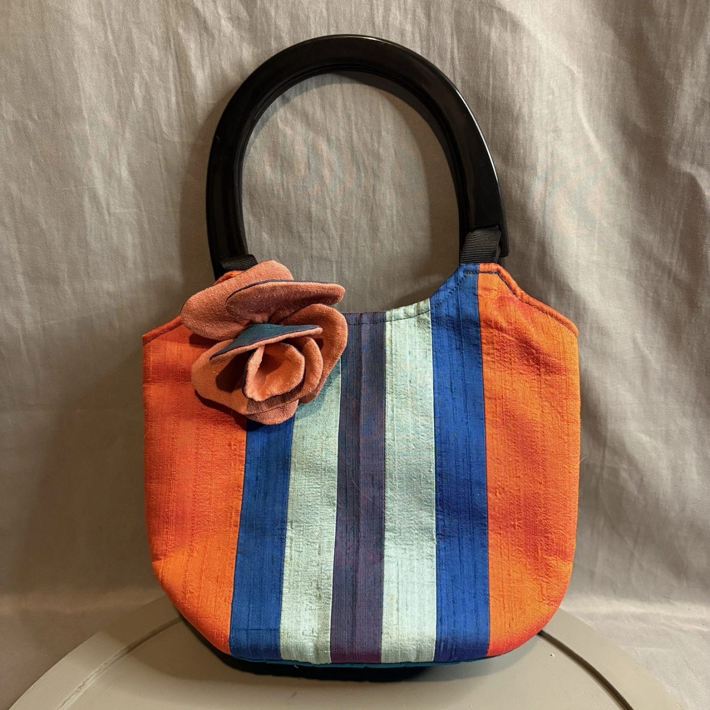 Handwoven raw silk purse with black handles, blue and orange vertical stripes, and a large beige flower on top. Handwoven raw silk purse with black handles, blue and orange vertical stripes, and a large beige flower on top.