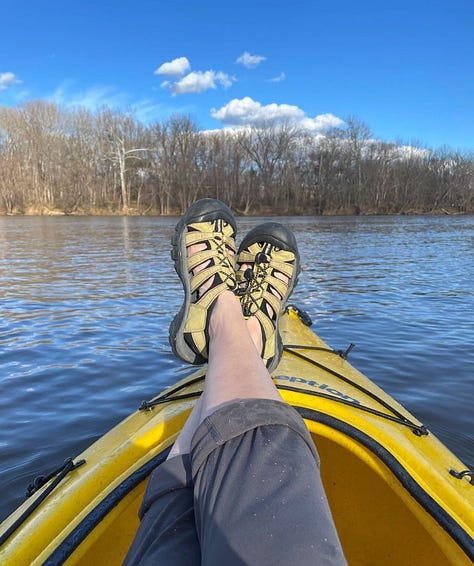 Feet in sandals resting on edge of kayak, pants rolled up. Photo of front of yellow kayak in river. Feet resting on windowsill in fleece socks, pants rolled down.