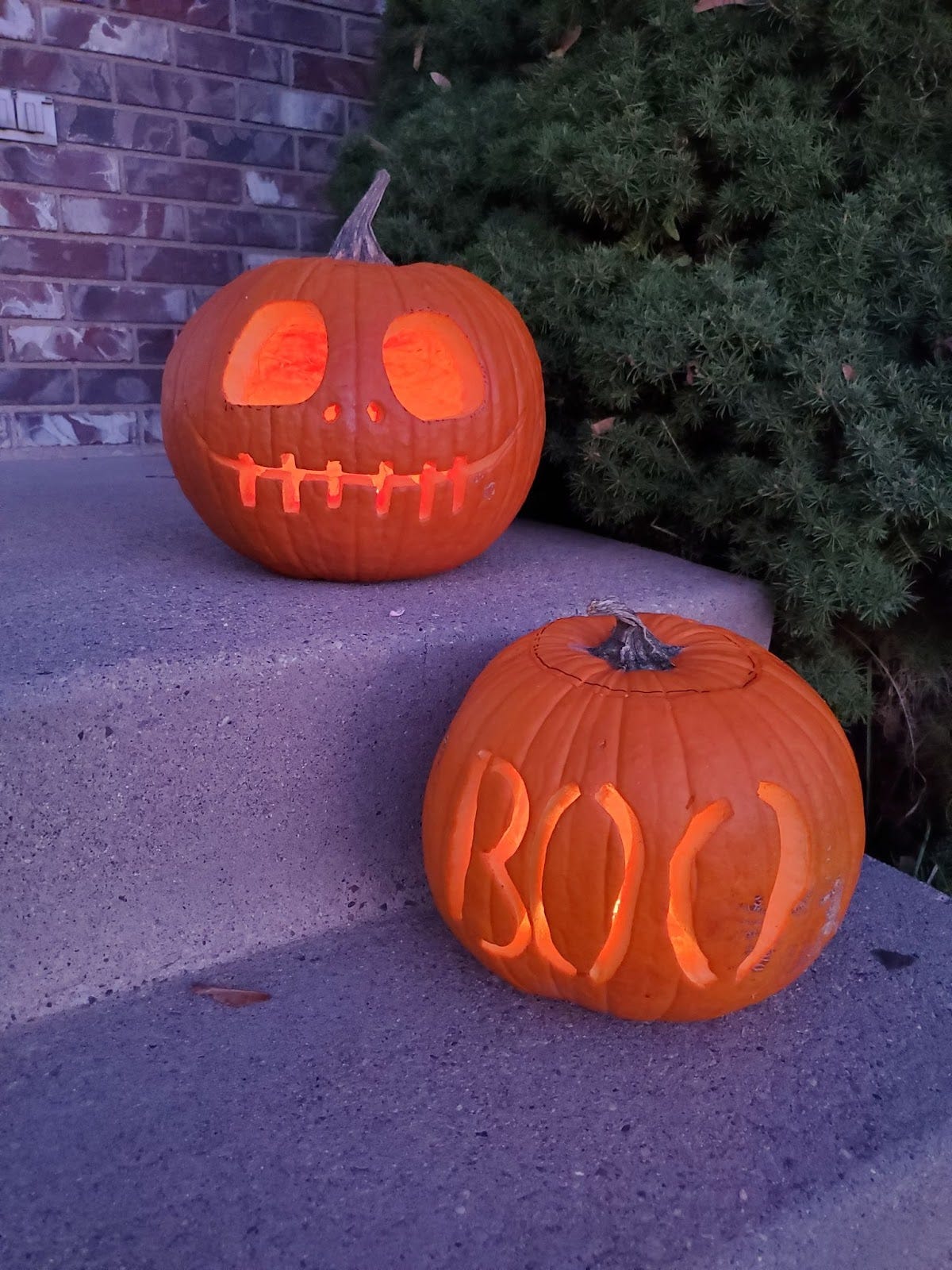 Two jack-o-lanterns on a set of concrete steps to a front porch. One is Jack Skellington and the other is carved to say “Boo” Two jack-o-lanterns on a set of concrete steps to a front porch. One is Jack Skellington and the other is carved to say “Boo”