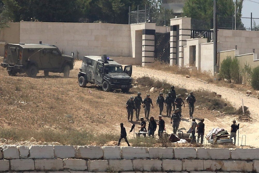 IOF personnel stand guard next to settlers in front of Palestinian homes that they have blockaded in the village of Qusra, south of Nablus, in the occupied West Bank, Wednesday, Aug. 12, 2026. (AP Photo/Nidal Eshtayeh) IOF personnel stand guard next to settlers in front of Palestinian homes that they have blockaded in the village of Qusra, south of Nablus, in the occupied West Bank, Wednesday, Aug. 12, 2026. (AP Photo/Nidal Eshtayeh)