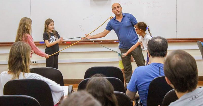 Um homem e três crianças mexendo em uma corda na frente de uma sala de aula com o público assistindo