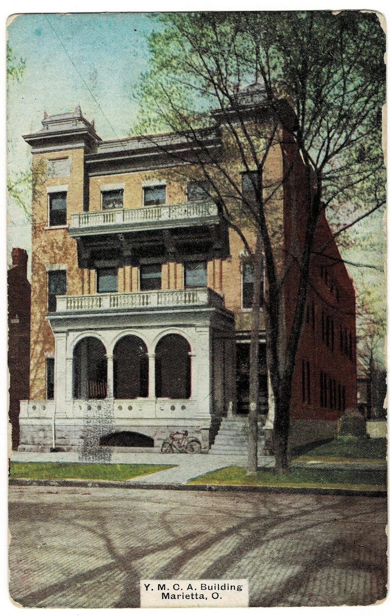 May include: Vintage postcard featuring the Y.M.C.A. Building in Marietta, Ohio. The multi-story building has a light brown facade with balconies and arched entryways. A bicycle is parked near the entrance. The postcard is aged with a faded appearance.