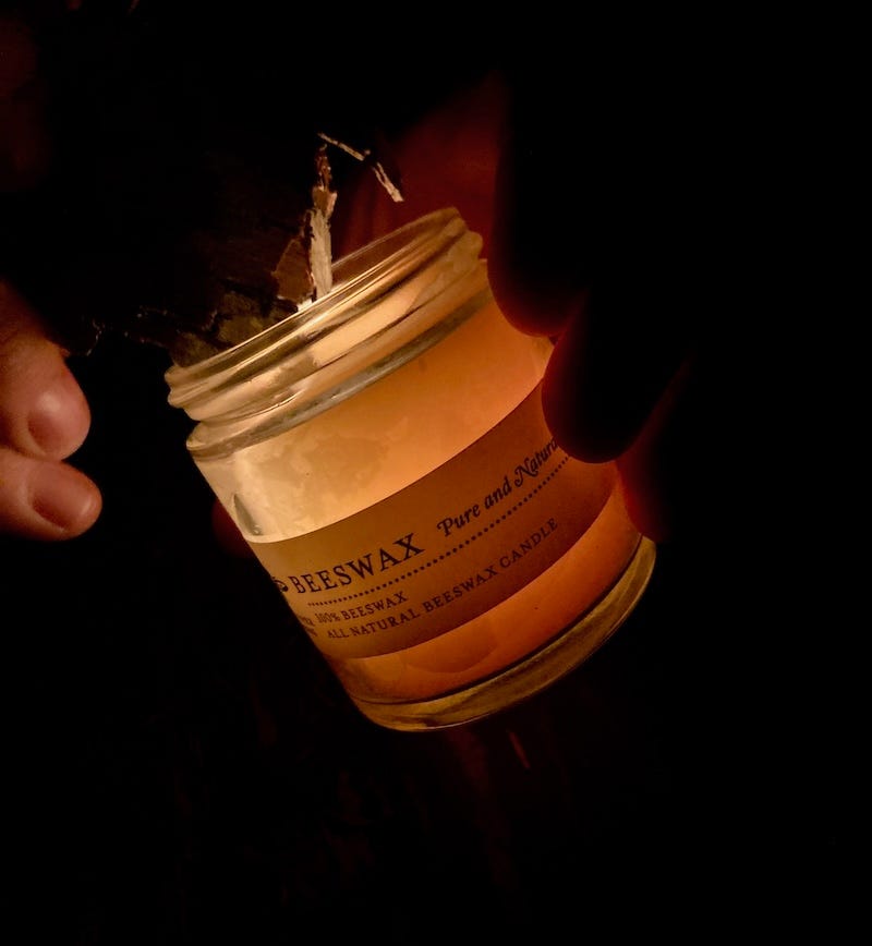 a person is holding a jar that is glowing because it contains a beeswax candle. the person is using the candle to light a piece of birch bark.