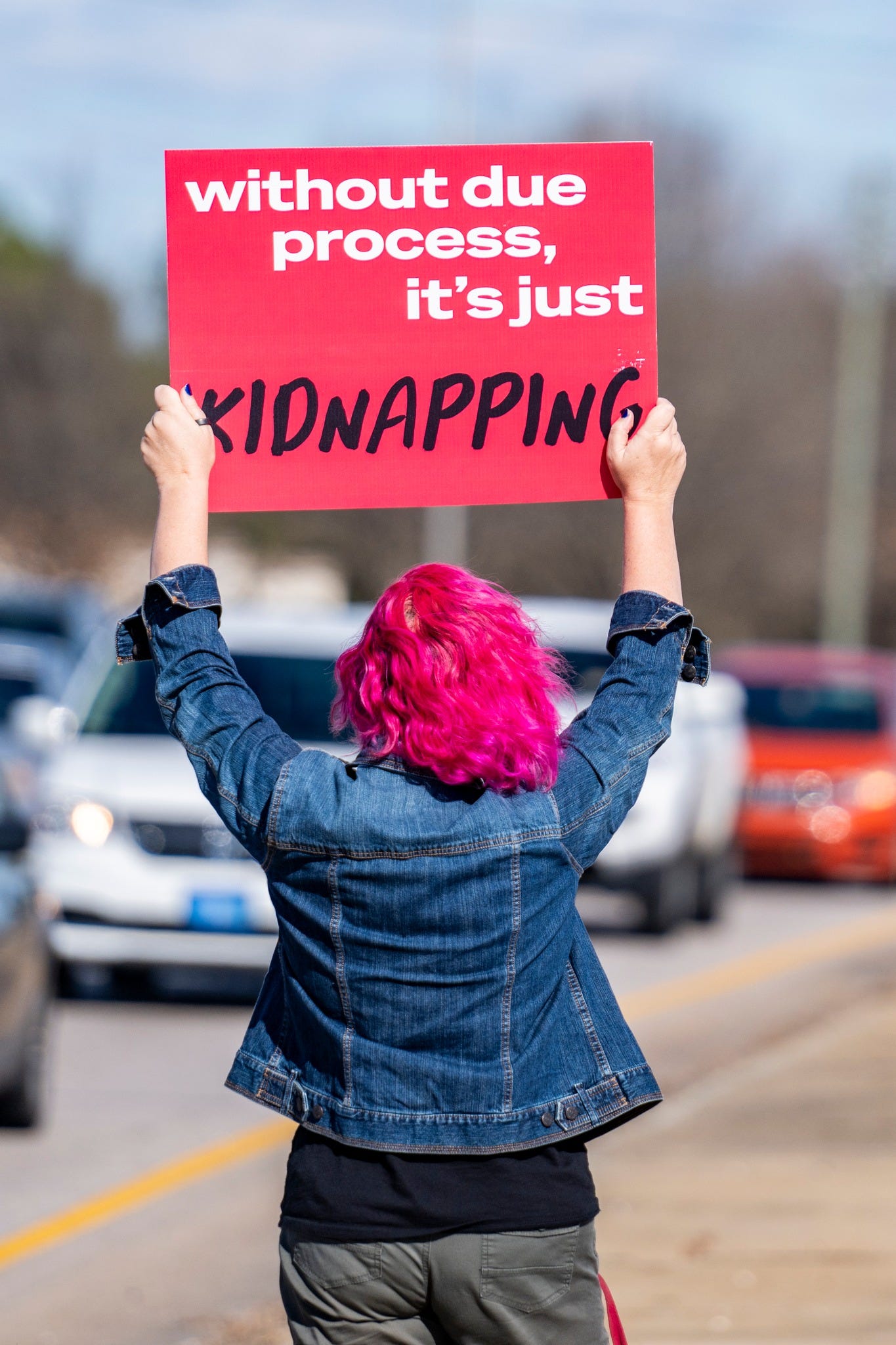 Person with bright pink hair holding a red protest sign that reads, “without due process, it’s just KIDNAPPING.”