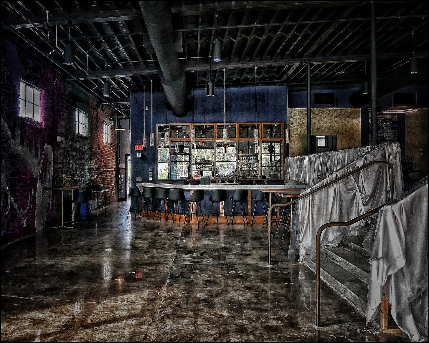 The interior of what appears to be a bar or nightclub -- either abandoned, or headed in that direction. Ceiling fixtures -- pipes, ductwork, electrical conduits -- are exposed; one wall, with small, high windows, is of exposed brick. The bar itself is a simple arc, like a portion of a circle, and there are about 10 barstools lined up before it. The wall behind the bar is mirrored; the shelves are bare. Various furniture and other fixtures are draped in clear plastic.