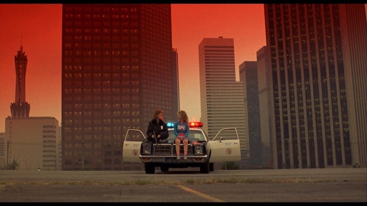 Catherine Mary Stewart and Kelli Maroney sit on a police car under a red-tinted sky in a deserted Los Angeles, from Night of the Comet (1984).