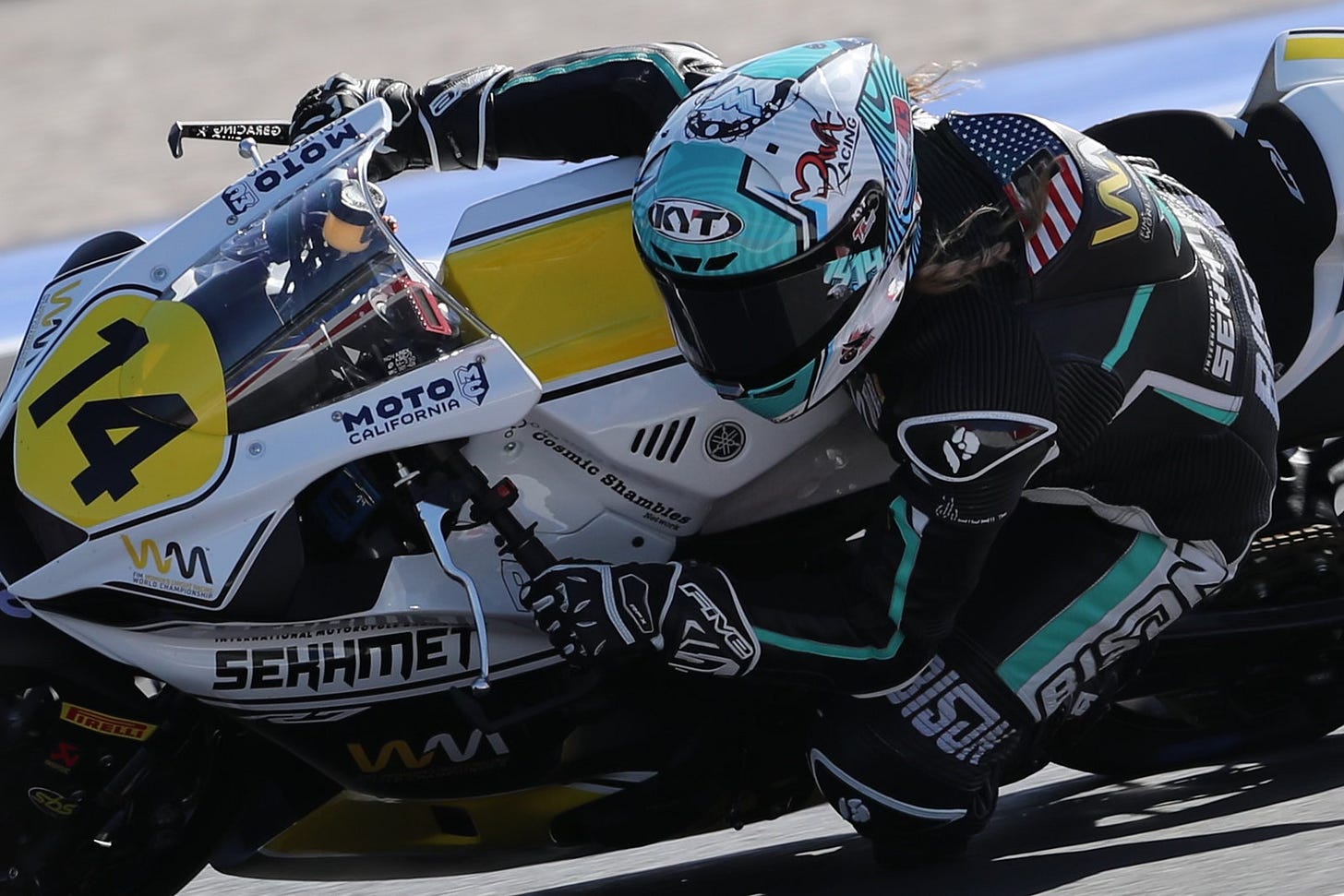 A white and black motorcycle with the Number 14 and sponsorship from Cosmic Shambles and Moto California goes through a turn