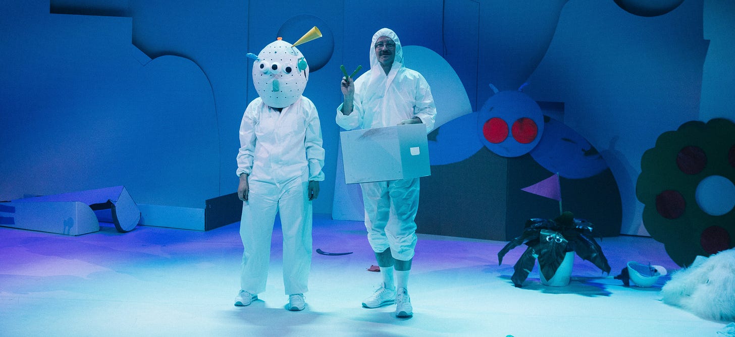 Two figures dressed in white hazmat suits, stand in the middle of a blue stage set filled with assorted cutouts of shapes, plants and animals. One is pulling items from a cardboard box, the other has their hands by their sides and is wearing an alien mask.