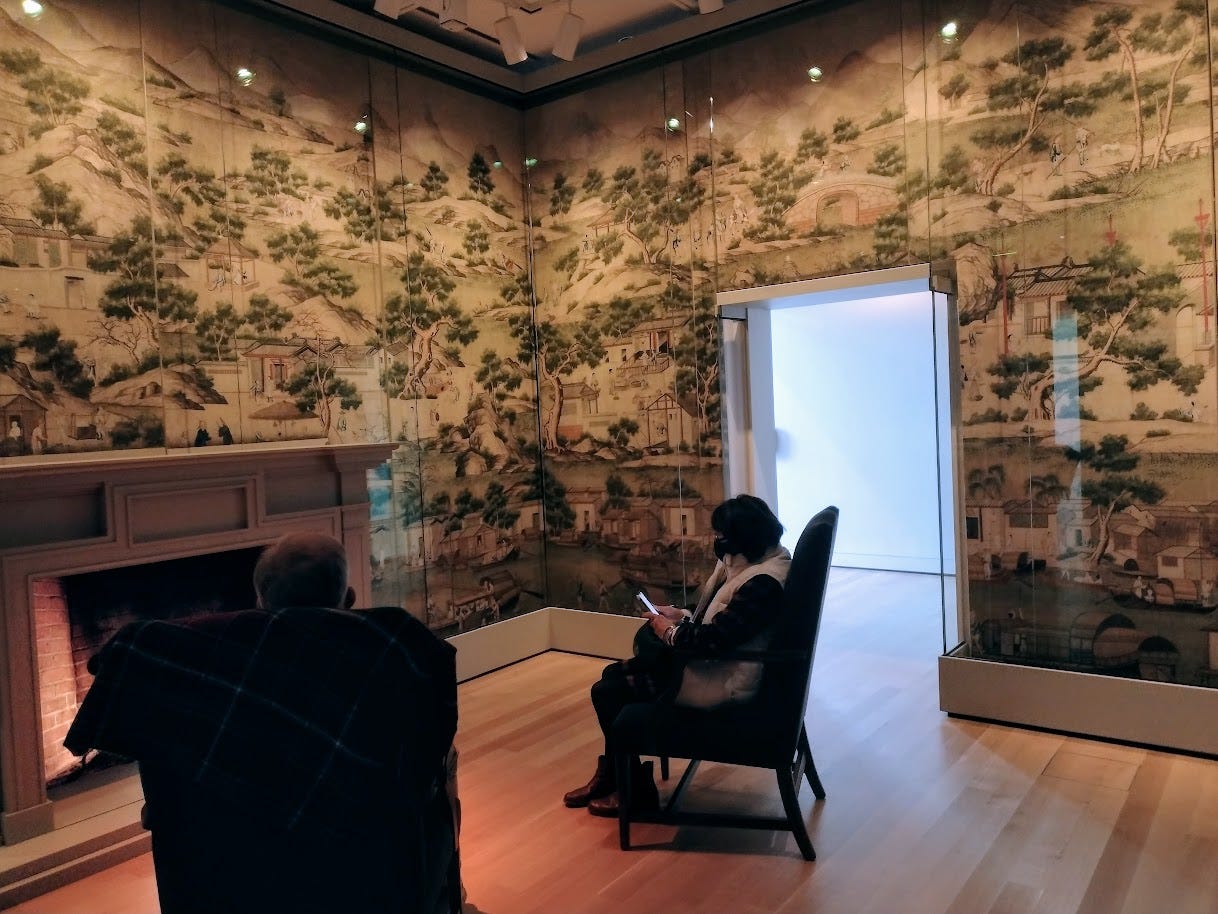 Man and woman in armchairs before fireplace, surrounded by wallpaper with drawings from China
