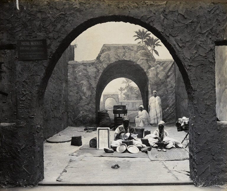 Four Nigerian men in traditional dress are seated surrounded by items featuring in a metalwork display.
