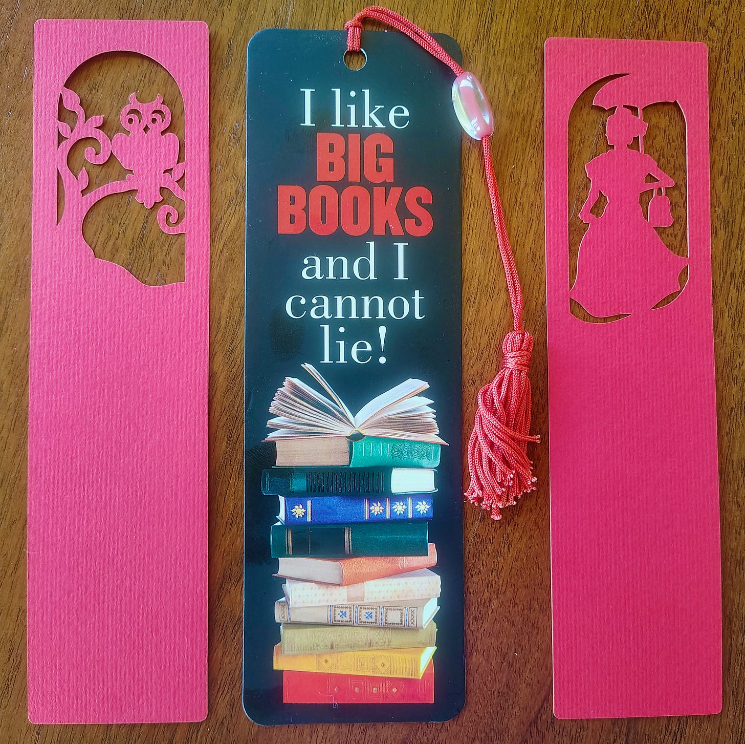 three bookmarks lying vertically next to each other with brown wood background. the ones on the right and left are both red with cut outs at the top, the left one an owl, the right one a vintage headshot of a woman. The middle bookmark is wider than the others and shows a stack of books and the letters I Like Big Books! It has a red tassle. 