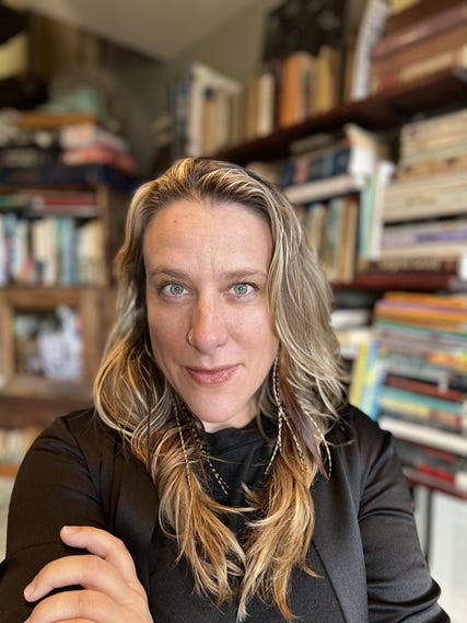 Photograph of a white woman with a beguiling smile in front of stacks of books in the background.