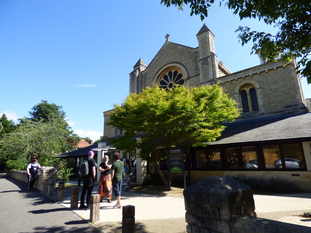 A group of Hutchmoot attendees standing around in the sunshine outside St Andrews church in Oxford.
