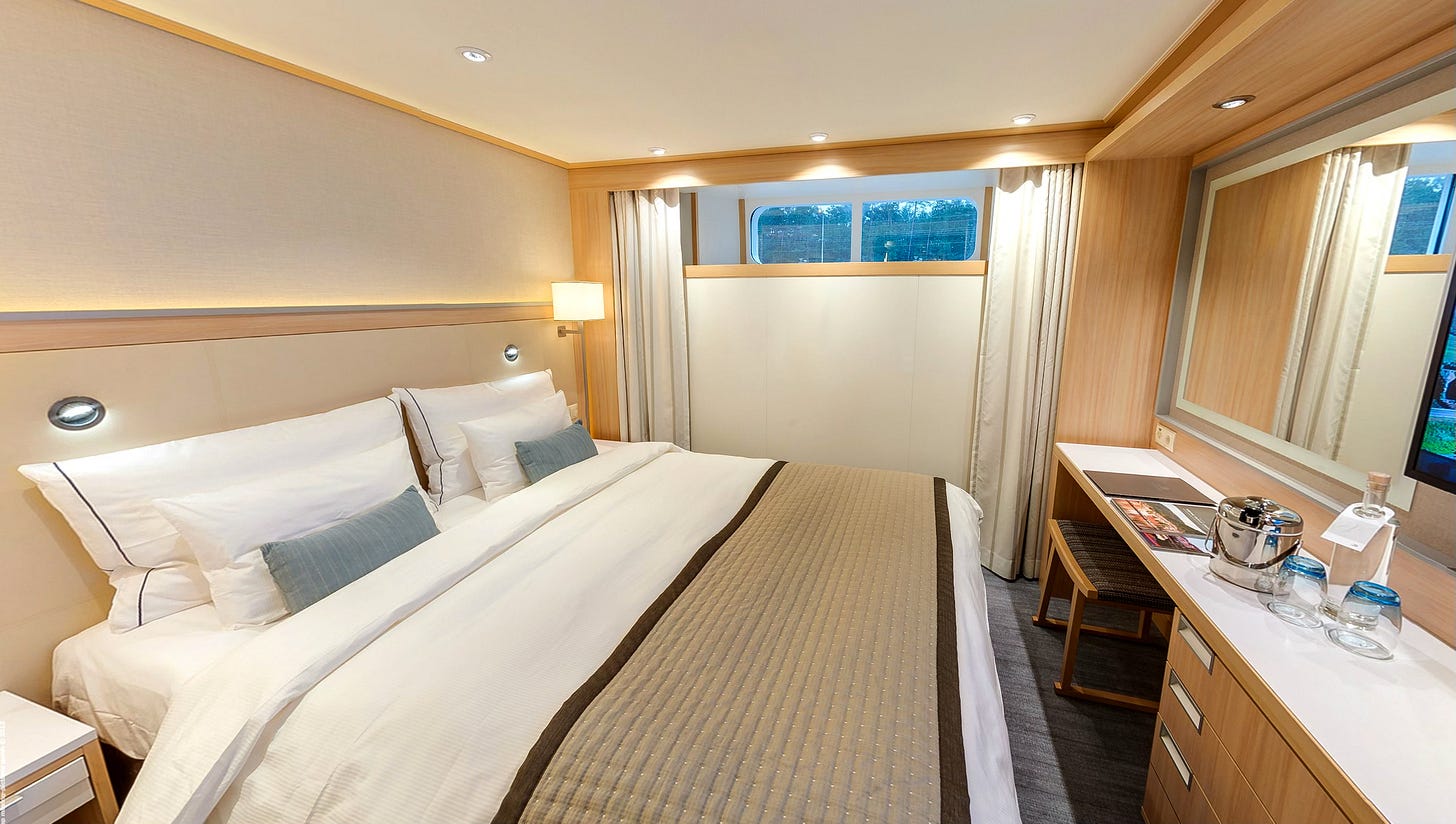 A standard stateroom on a Viking river cruise, featuring a queen-size bed, a small window, a desk area, and neutral decor. A standard stateroom on a Viking river cruise, featuring a queen-size bed, a small window, a desk area, and neutral decor.
