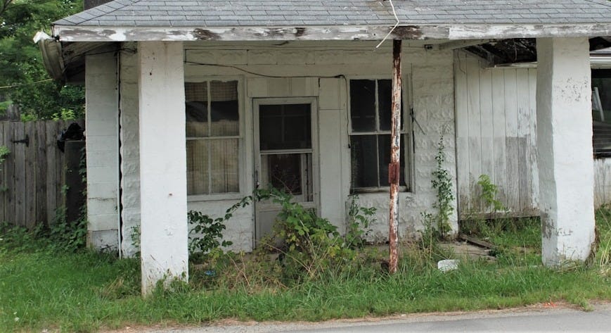A derelict white clapboard house with a sagging porch roof, peeling paint and weeds growing across the front A derelict white clapboard house with a sagging porch roof, peeling paint and weeds growing across the front