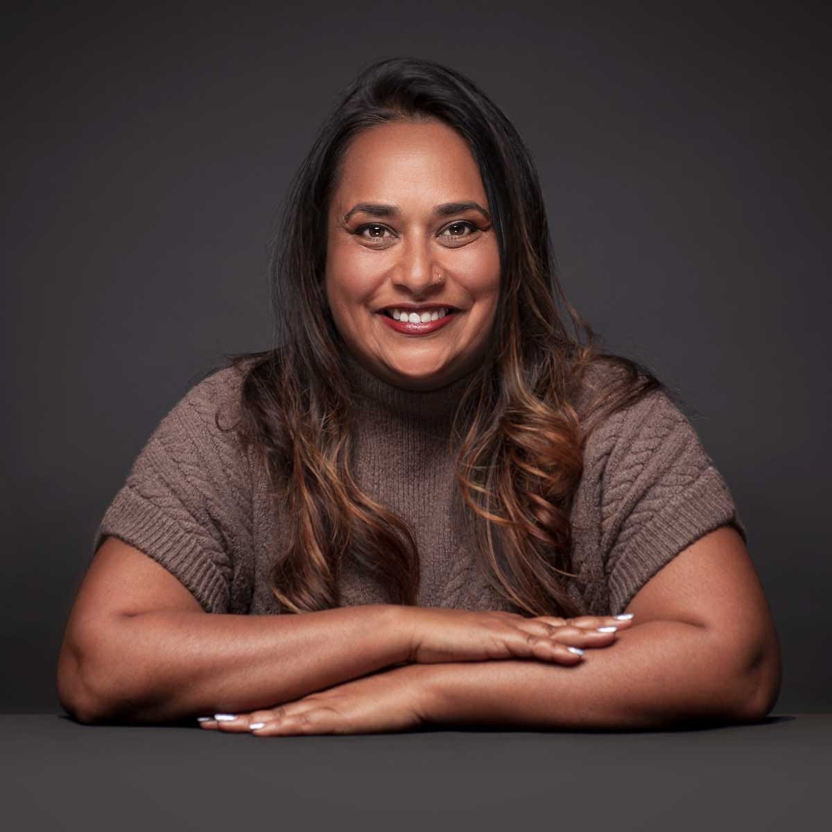 A headshot photo of Vini, looking straight at the camera, smiling big, wearing a brown short sleeved sweater. She has brown skin, long brown hair and her hands are crossed in front of her.