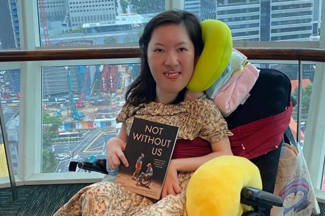 A Singaporean femme powerchair-user poses with an anthology called Not Without Us against floor to ceiling windows that show a city far below.