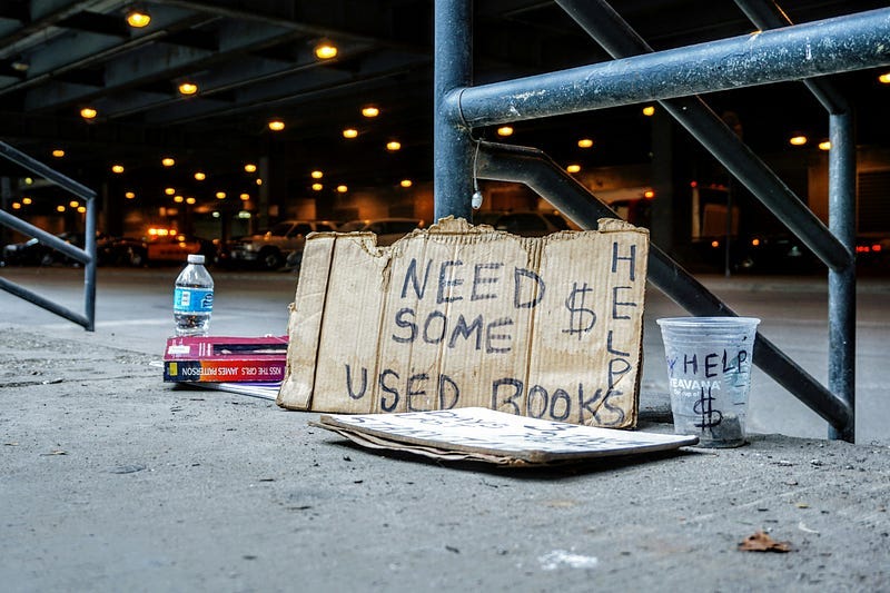 A sign reads, “Need some money, used books,” next to a cup for change.