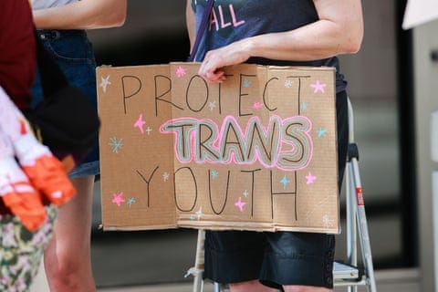 close-up of person holding cardboard sign saying 'protect trans youth' outlined in pink and blue