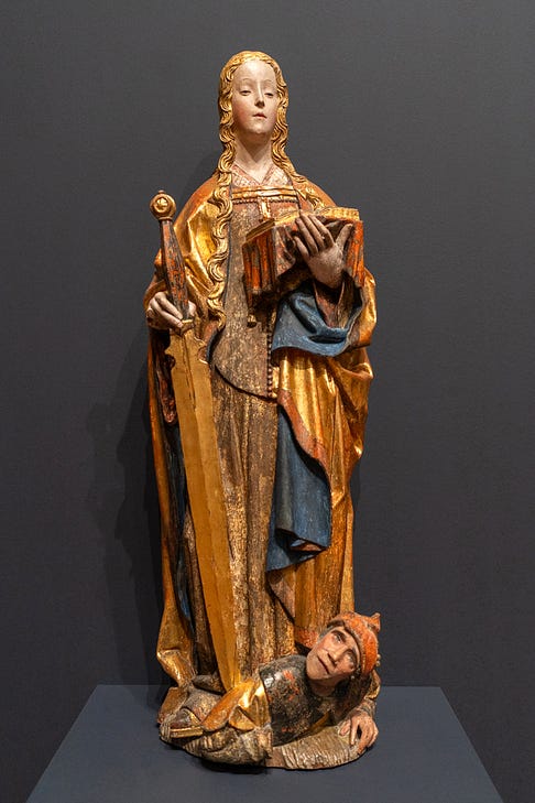 a medieval polychrome wood statue of St Catherine of Alexandria. She wears blue and gold robes and is holding a sword in one hand and a book in the other