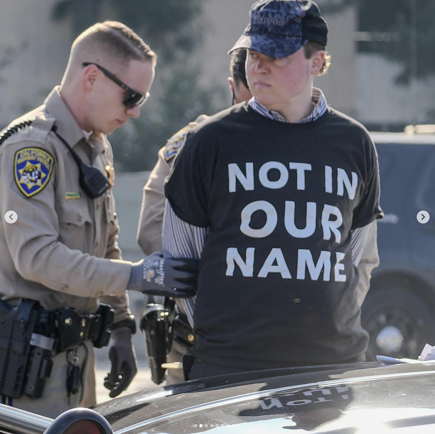 A protester wearing a shirt that says "NOT IN OUR NAME" being detained, possibly arrested by 2 California officers