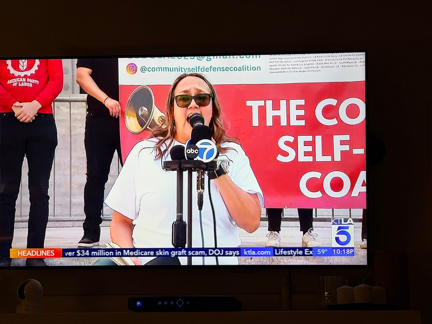 an image from a televsion evening newscast. A light skinned Puerto Rican woman with dark hair and sunglasses stands holding a mic and speaking in front of verious television news station mics. She's wearing a white t shirt. an image from a televsion evening newscast. A light skinned Puerto Rican woman with dark hair and sunglasses stands holding a mic and speaking in front of verious television news station mics. She's wearing a white t shirt.