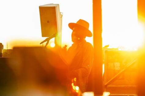 A gallery of three photos of Gary Clark Jr. and his band performing on an outdoor stage. The left photo shows him in silhouette against a bright orange sun, the middle photo shows him playing guitar, and the right photo shows him playing next to his bassist.